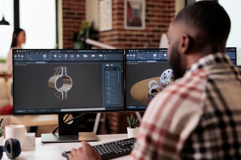 Person working at a desk with two monitors displaying 3D CAD models of mechanical metal parts.