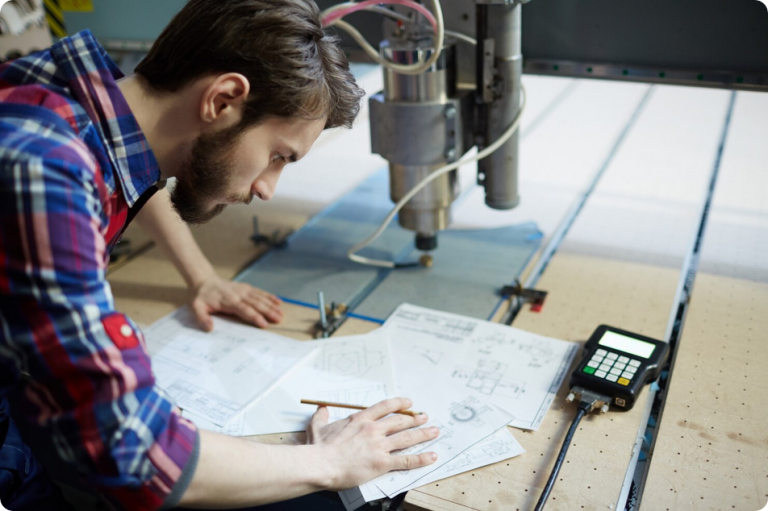 A trainee studying technical drawings beside a CNC router machine during a CNC machining training session in Mississauga.